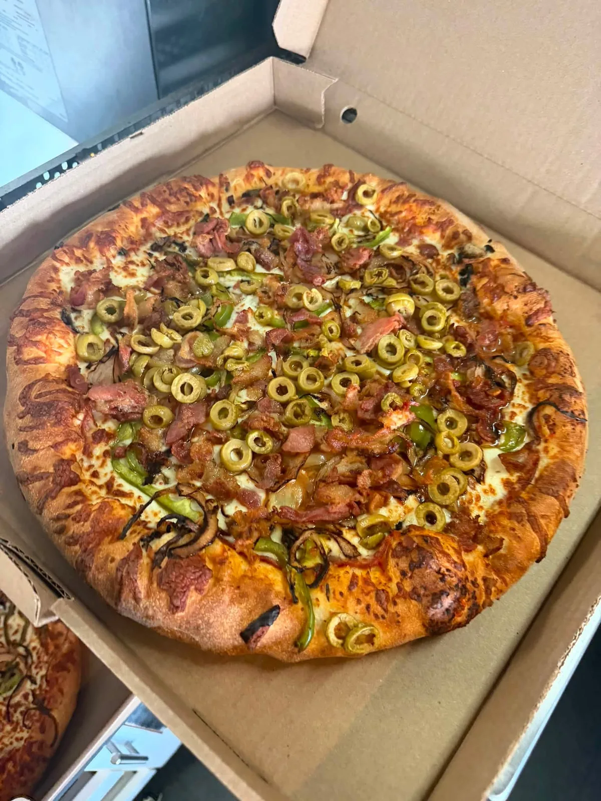 Pizza garnie de bacon, d’olives vertes, de poivrons et d’oignons, dans sa boîte.